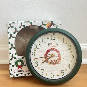 May include: A round wall clock with a dark green frame and a white face featuring black numbers, gold bell icons, and a central Christmas wreath illustration. The text Merry Christmas is printed above the wreath, with Quartz below it.