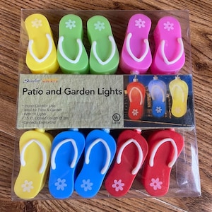 May include: A set of 10 flip-flop shaped patio and garden lights in yellow, green, pink, blue, and red. Each light features a white strap and a small white flower detail. The packaging displays the text Sienna Nights Patio and Garden Lights.