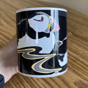 May include: A white ceramic mug featuring a graphic illustration of a puffin with a black, white, and grey body and a colorful beak. The design includes a stylized yellow and grey wave pattern against a solid black background on the mug surface.