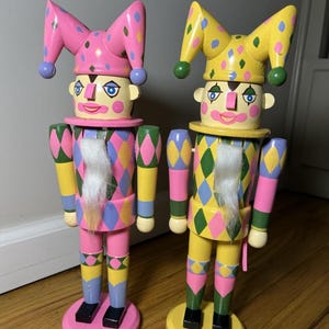 May include: Two wooden nutcracker figures dressed as jesters, each wearing a multi-pointed hat with pom-poms. One figure features a pink color scheme, while the other is primarily yellow. Both have harlequin-patterned outfits, white beards, and painted facial features, standing on circular bases.
