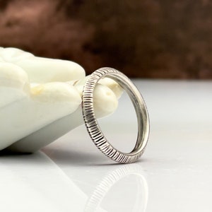 May include: A silver ring with a textured, ridged design. The ring is round and appears to be made of metal. The ridges run around the entire band of the ring. The ring is displayed on a white surface.