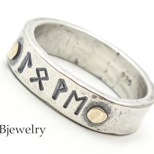 May include: A silver band ring with a hammered finish and black engraved runes. The ring has two small gold dots on the band. The ring is marked 925.