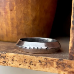 May include: A silver ring with a textured band. The ring is sitting on a wooden surface.