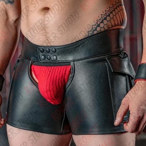 Puede incluir: Pantalones cortos de cuero negro con una cintura ancha de seis botones a presión, una abertura central que revela una bolsa de tela roja acanalada y bolsillos laterales. Se combina con muñequeras de cuero negro a juego con forro rojo.