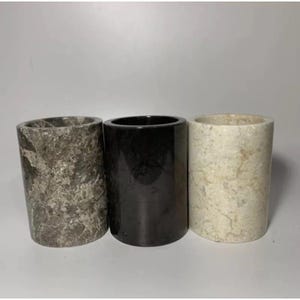 Può includere: Tre contenitori cilindrici in pietra disposti in fila. Il contenitore a sinistra è grigio con venature bianche, quello centrale è nero solido con finitura lucida e quello a destra è color crema con sottili venature beige.