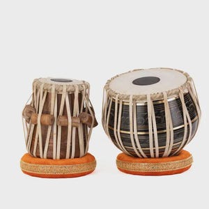 May include: A pair of tabla drums featuring a smaller wooden drum and a larger metallic drum with black and gold horizontal bands. Both drums have white drumheads, tension ropes, and are resting on circular orange fabric cushions with gold trim.