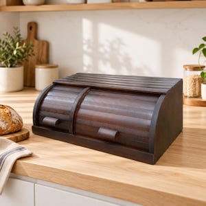 May include: A dark brown wooden bread box featuring a roll-top lid with two horizontal handles. The rectangular storage container has a slatted design and is positioned on a light-colored wooden countertop surface.