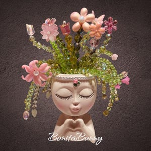 Peut inclure: Un pot de fleurs en céramique crème en forme de tête humaine avec les yeux fermés et les mains formant un cœur. Le pot est orné d'un arrangement décoratif de perles de cristal vertes et de diverses perles en forme de fleurs roses et pêche.