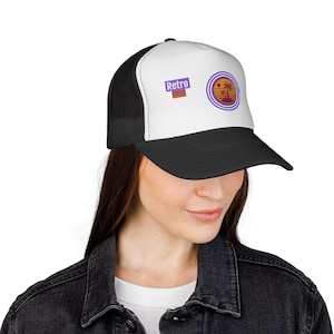 Puede incluir: Una gorra de camionero negra y blanca con un panel frontal blanco y un estampado gráfico. El estampado incluye el texto Retro Vibes en morado y naranja, junto a un diseño circular con anillos morados, un sol amarillo y una palmera negra.