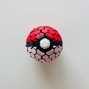 May include: A spherical object featuring a red top half and a white bottom half, both composed of a textured, interlocking hexagonal pattern. A black band separates the two halves, and a circular white button is centered on the front.