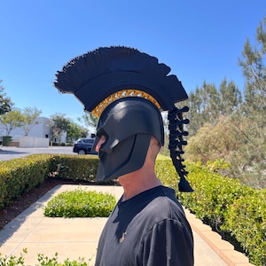 May include: A matte black Spartan-style helmet featuring a prominent crest with a textured black top and a gold-coloured interior detail. The helmet includes a spinal column-shaped extension trailing down the back of the head piece.