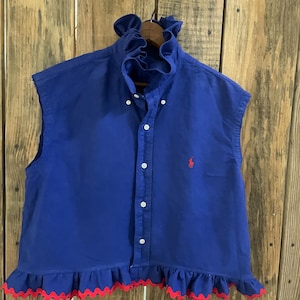May include: A sleeveless royal blue button-down shirt featuring a high ruffled collar and a ruffled hemline with a red scalloped edge. The shirt includes a small red embroidered logo on the left chest and white buttons down the front placket.