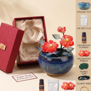 May include: A DIY craft kit featuring a dark blue ceramic pot, red artificial flowers with pearl centres, air-dry clay, a small bottle of essential oil, green decorative pebbles, floral tape, and florist wire, all presented with a red gift box.
