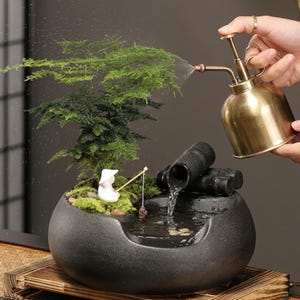 Könnte beinhalten: Ein runder schwarzer Keramik-Zimmerbrunnen mit einer kleinen grünen Pflanze, einer weißen Angler-Figur und einem schwarzen Auslauf in Bambusoptik. Ein messingfarbener Metallzerstäuber sprüht Wasser auf die Pflanze über dem Brunnenbecken.