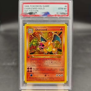 May include: A 1999 Pokemon Game Charizard-Holo 1st Edition trading card, graded GEM MT 10, encased in a clear protective plastic slab. The card features a fiery orange dragon illustration, yellow border, and text detailing its attacks and stats.