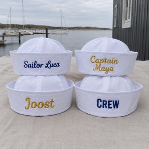 Może przedstawiać: Cztery białe czapki marynarskie ułożone w siatkę dwa na dwa. Każda czapka posiada wyhaftowany tekst: w lewym górnym rogu Sailor Luca na niebiesko, w prawym górnym rogu Captain Maya na złoto, w lewym dolnym rogu Joost na złoto, a w prawym dolnym rogu CREW na niebiesko.