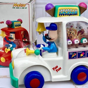May include: A white plastic toy ice cream truck with blue wheels, a red center hub, and a driver figure. The roof features two ice cream cone decorations and a sign reading ICECREAM. The side displays a menu with images of treats and the text YUM YUM.