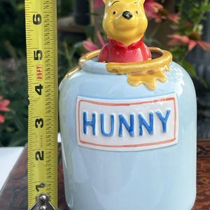 May include: A light blue ceramic jar featuring a yellow Winnie the Pooh figure emerging from the top. The jar has a yellow honey drip design and a rectangular label with the word HUNNY in blue letters. A yellow measuring tape shows a height of 6 inches.