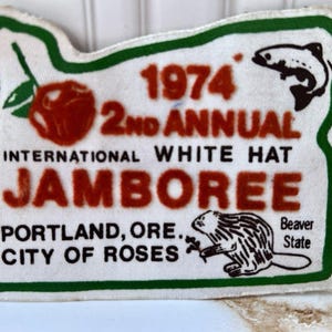 May include: A white fabric patch shaped like the state of Oregon with a green border. It features a red rose, a black fish, and a black beaver illustration. Text reads: 1974 2ND ANNUAL INTERNATIONAL WHITE HAT JAMBOREE PORTLAND, ORE. CITY OF ROSES Beaver State.