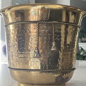 May include: A polished brass planter featuring a hammered texture, a wide flared rim, and two horizontal rope-style bands. Two decorative hanging tassels are attached to the upper rope band, and the base shows minor surface wear and discoloration.
