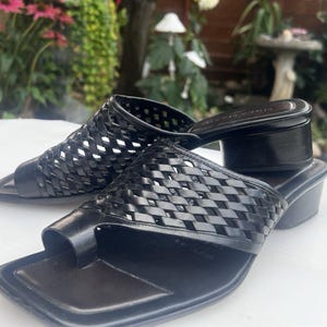 May include: A pair of black leather sandals featuring a woven lattice upper design, a square toe, and a low block heel. The sandals include a toe loop strap and a smooth footbed, providing a structured look with a consistent dark finish throughout.
