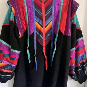 May include: A long-sleeved black jacket featuring vibrant, multi-colored fabric panels in shades of red, purple, blue, and teal. The back displays a chevron-patterned central panel accented with hanging ribbons and intricate geometric patchwork.