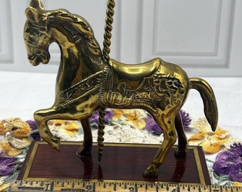 May include: A brass carousel horse figurine with a twisted pole, mounted on a rectangular wooden base. A wooden ruler placed in front shows the base measures approximately 5.5 inches in length and the horse stands about 5 inches tall.