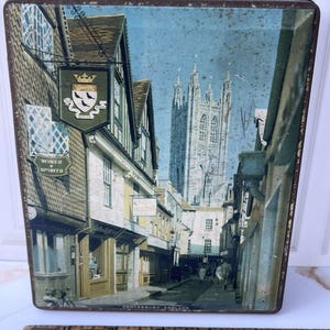 May include: A rectangular metal tin featuring a printed scene of a street in Canterbury, England, with a cathedral in the background. A wooden ruler is placed in front of the tin, showing a width measurement of approximately 7.5 inches.