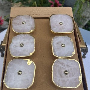 May include: A set of six square drawer knobs featuring translucent pale pink rose quartz stone faces with irregular, textured gold-toned metallic edges. Each knob has a small, centered, circular metallic stud and is arranged in a cardboard box.