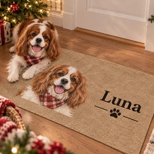 May include: A rectangular tan doormat featuring a printed portrait of a brown and white Cavalier King Charles Spaniel wearing a red and white plaid bandana. The name Luna is printed in black text above a small black paw print graphic.