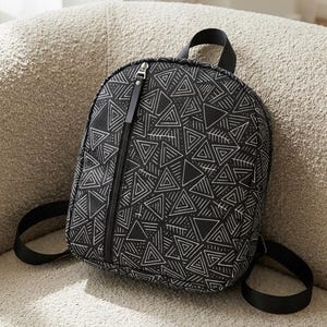Puede incluir: Una mochila negra con un patrón geométrico blanco de triángulos y líneas. La bolsa tiene una cremallera frontal vertical con un tirador negro, un asa superior y correas de hombro negras ajustables. El diseño presenta formas triangulares.