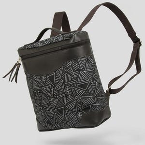 Puede incluir: Una mochila negra con un patrón geométrico de triángulos blancos. La bolsa tiene un asa superior marrón oscuro, un cierre de cremallera con dos tiradores y correas ajustables de color marrón. La base y el borde superior son de color marrón.