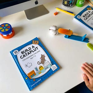 May include: A blue activity book titled Build A Catapult STEM Engineering Challenge Ages 7-12 with a cartoon cat illustration. Nearby are a blue lolly stick catapult, an orange lolly, a cotton ball, a yellow pencil, and a toy.