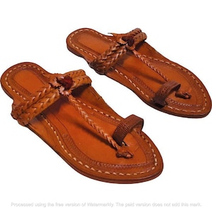 May include: A pair of tan leather sandals featuring braided straps across the top, a central vertical braided strap, and a toe loop. The edges of the soles are decorated with a stitched pattern, and the design includes a small dark brown knot detail.