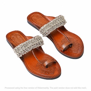 May include: A pair of brown leather sandals featuring a wide strap adorned with silver-toned metallic beads and a toe loop. The footbed is embossed with a floral pattern, and the design includes a braided leather detail connecting the strap to the toe.