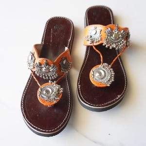 May include: A pair of flat sandals featuring dark brown footbeds and orange leather straps. The straps are adorned with intricate silver-toned metal embellishments and small dangling charms that extend to a circular toe post detail.