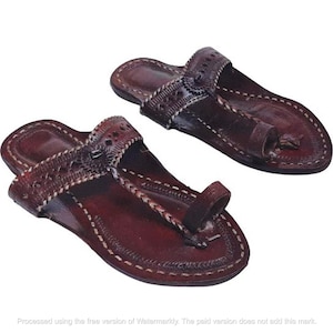 May include: A pair of dark brown leather sandals featuring a toe loop design, intricate cut-out patterns on the wide strap, and decorative braided detailing. The edges of the soles and straps are finished with visible white stitching throughout.