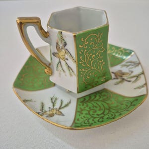 Puede incluir: Una taza de té hexagonal de porcelana con su plato a juego, que presenta una base blanca con paneles verdes y detalles de filigrana dorada. El conjunto está decorado con ilustraciones pintadas a mano de pequeños pájaros sobre ramas.