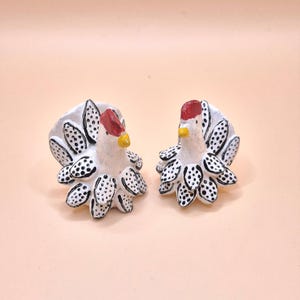 May include: Two small, hand-painted ceramic chicken figurines featuring white bodies with black polka-dot patterns on their feathers. Each chicken has a bright red comb on top of its head and a small yellow beak, standing in a side-by-side arrangement.