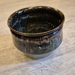 Può includere: Una piccola ciotola in ceramica rotonda caratterizzata da uno smalto scuro e maculato che sfuma da un nero profondo e lucido sul bordo a un grigio più chiaro e strutturato alla base. L'interno della ciotola presenta una finitura scura simile.