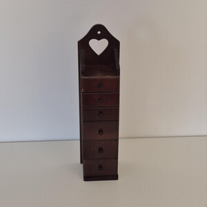 May include: A tall, dark brown wooden wall-mounted organizer featuring six stacked drawers with small black knobs. The top section includes an open compartment with a heart-shaped cutout and a pre-drilled hole for hanging on a wall surface.