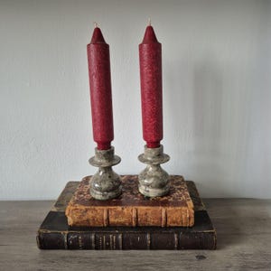 May include: Two tall, textured dark red taper candles are set into matching grey and white mottled stone candle holders. These holders are positioned on top of two stacked antique books with worn, weathered covers and visible spine text.