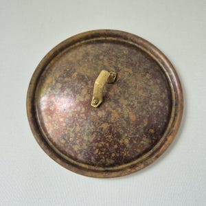 Könnte beinhalten: Ein runder Topfdeckel aus Kupfer mit einer verwitterten, fleckigen Patina mit dunkelbraunen und bronzefarbenen Oxidationsflecken. Der Deckel hat einen erhöhten Rand und einen mittigen, kantigen Messinggriff, der mit zwei Schrauben befestigt ist.