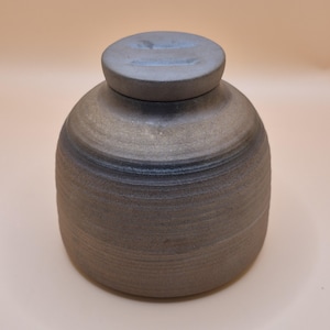 May include: A cylindrical ceramic jar with a matching lid, featuring a dark, matte, charcoal-gray finish with subtle horizontal ridges and a textured surface. The vessel has a wide, rounded body that tapers slightly toward the top opening.