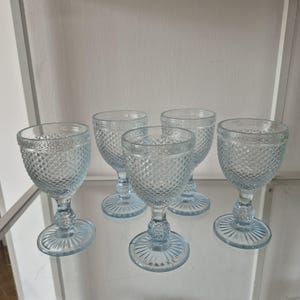 May include: A set of four light blue glass goblets featuring a textured diamond pattern on the bowl, a short stem with a decorative knob, and a circular base with a radial sunburst design. The glassware is translucent with a pale blue tint.