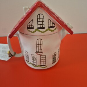 Peut inclure: Une théière en céramique blanche en forme de maison avec un toit rouge. La théière présente des fenêtres peintes en noir, des décorations de guirlandes vertes et une anse sur le côté gauche. Une petite étiquette blanche est attachée à l'anse.