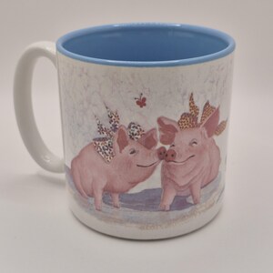 Op de afbeelding: Een witte keramische mok met een lichtblauwe binnenkant en handvat. De buitenkant is voorzien van een bedrukte illustratie van twee roze varkens met gestreepte strikken op hun hoofd, die samenstaan met een kleine rode vlinder erboven.