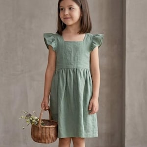 May include: A sage green dress featuring a square neckline, flutter sleeves, and a gathered waistline that falls to a knee-length skirt. The garment is constructed with a visible textured weave and is paired with a small, round, woven brown wicker basket.