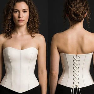 May include: A strapless white corset featuring a sweetheart neckline and vertical paneling. The back of the garment displays a lace-up closure with silver-toned metal eyelets and white cord lacing, providing a structured and fitted appearance.