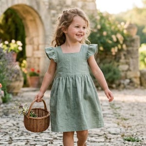 Könnte beinhalten: Ein salbeigrünes ärmelloses Kleid mit quadratischem Ausschnitt, gerüschten Schulterdetails und einem gerafften Rock, der über den Knien endet. Das Kleid besteht aus einem strukturierten Stoff und wird mit einem kleinen, runden, braunen Weidenkorb kombiniert.